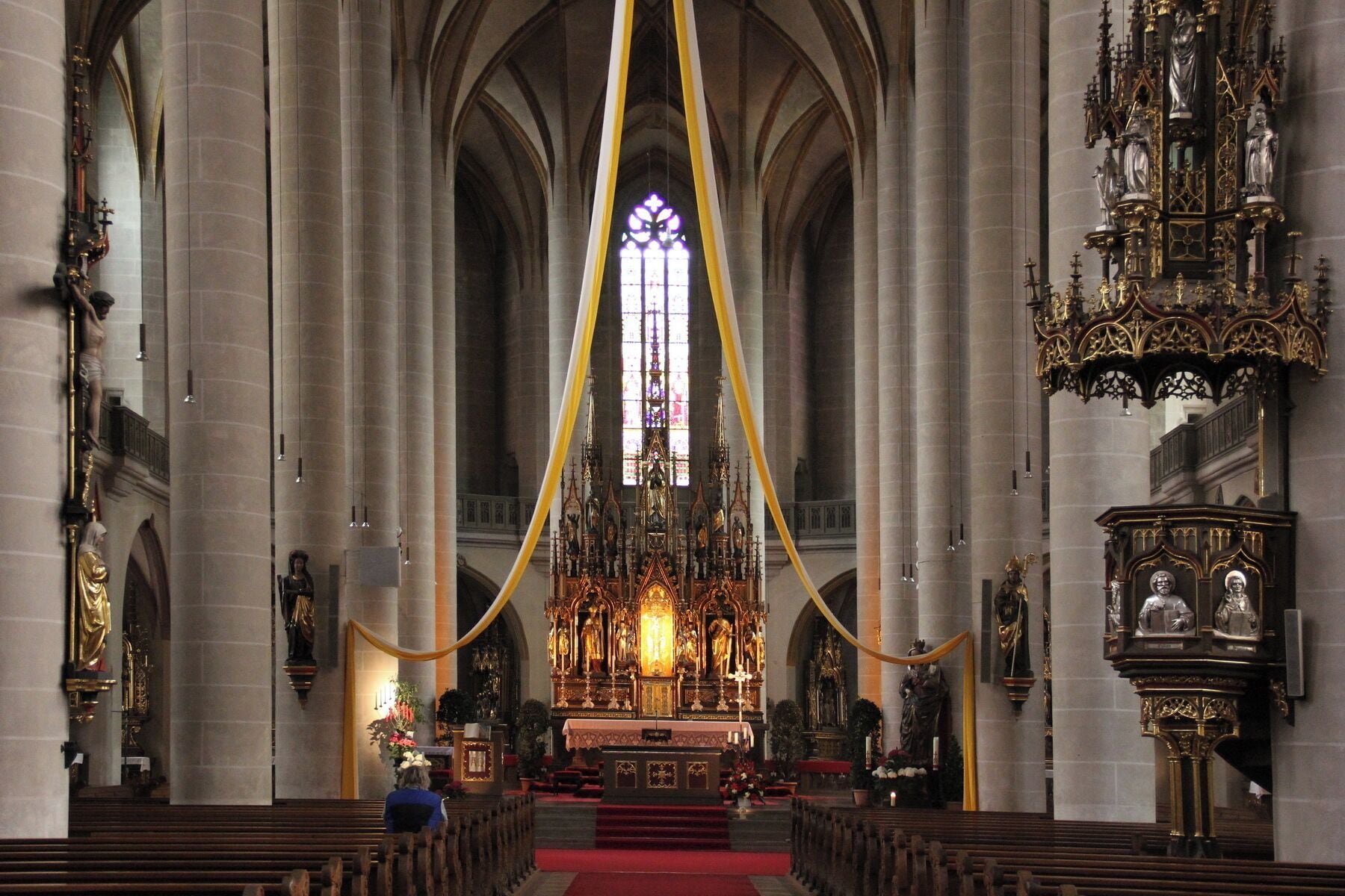 A grand cathedral interior with high vaulted ceilings, a central altar, and ornate religious statues.