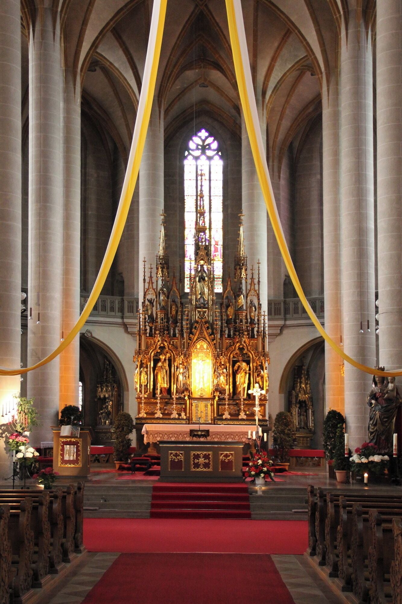 A cathedral interior with a central altar, high vaulted ceilings, and a large stained glass window.