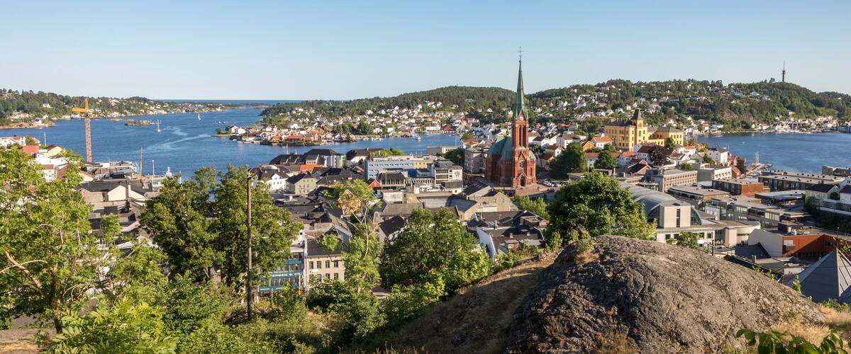 A coastal town with a prominent church spire, a harbor with boats, and a hilly landscape in the background.