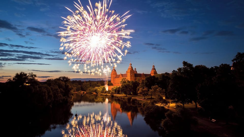 Un castillo junto a un lago, con reflejo de fuegos artificiales.