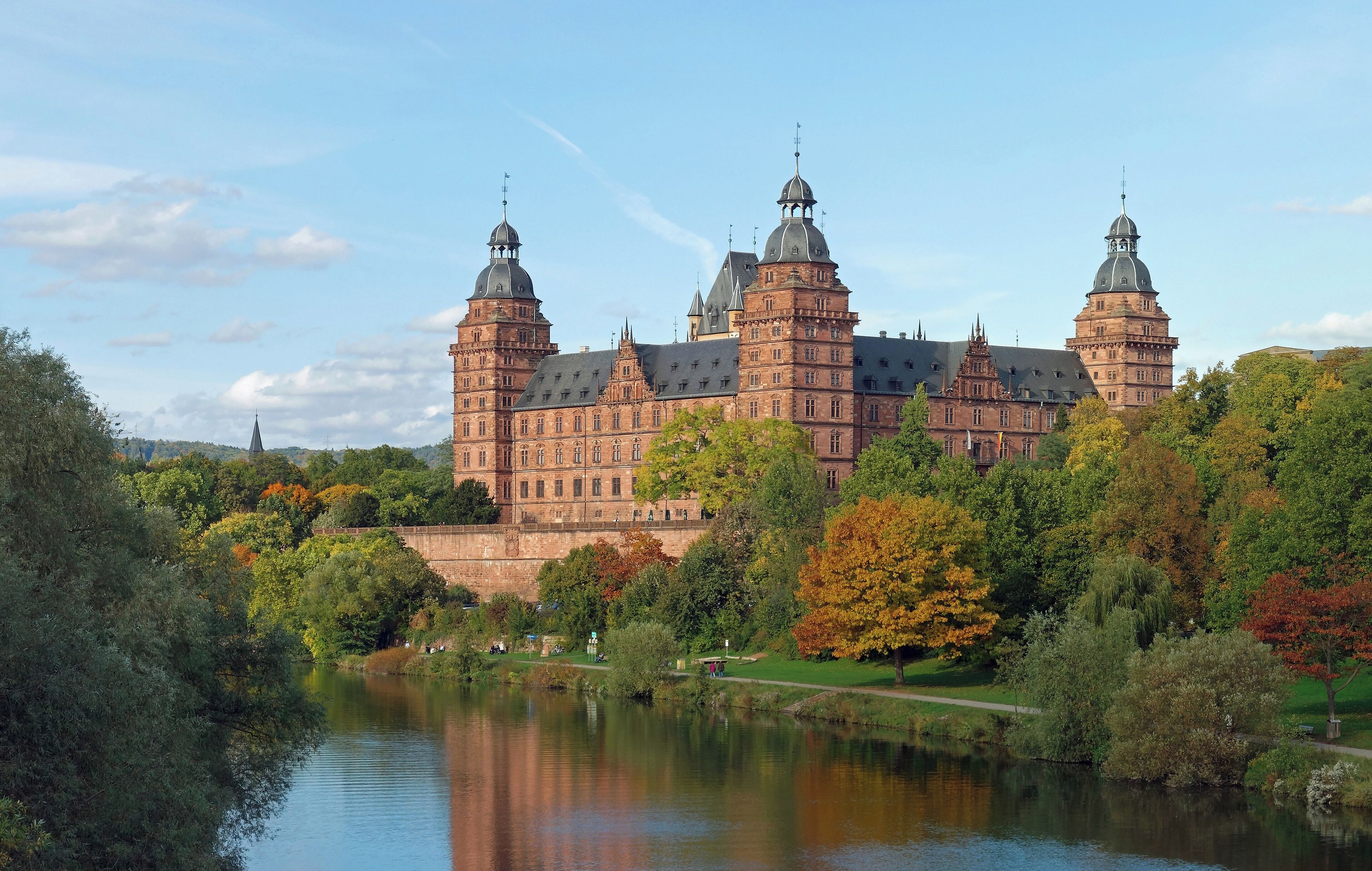 A historic building with multiple towers and domes, surrounded by a lake and autumn foliage.