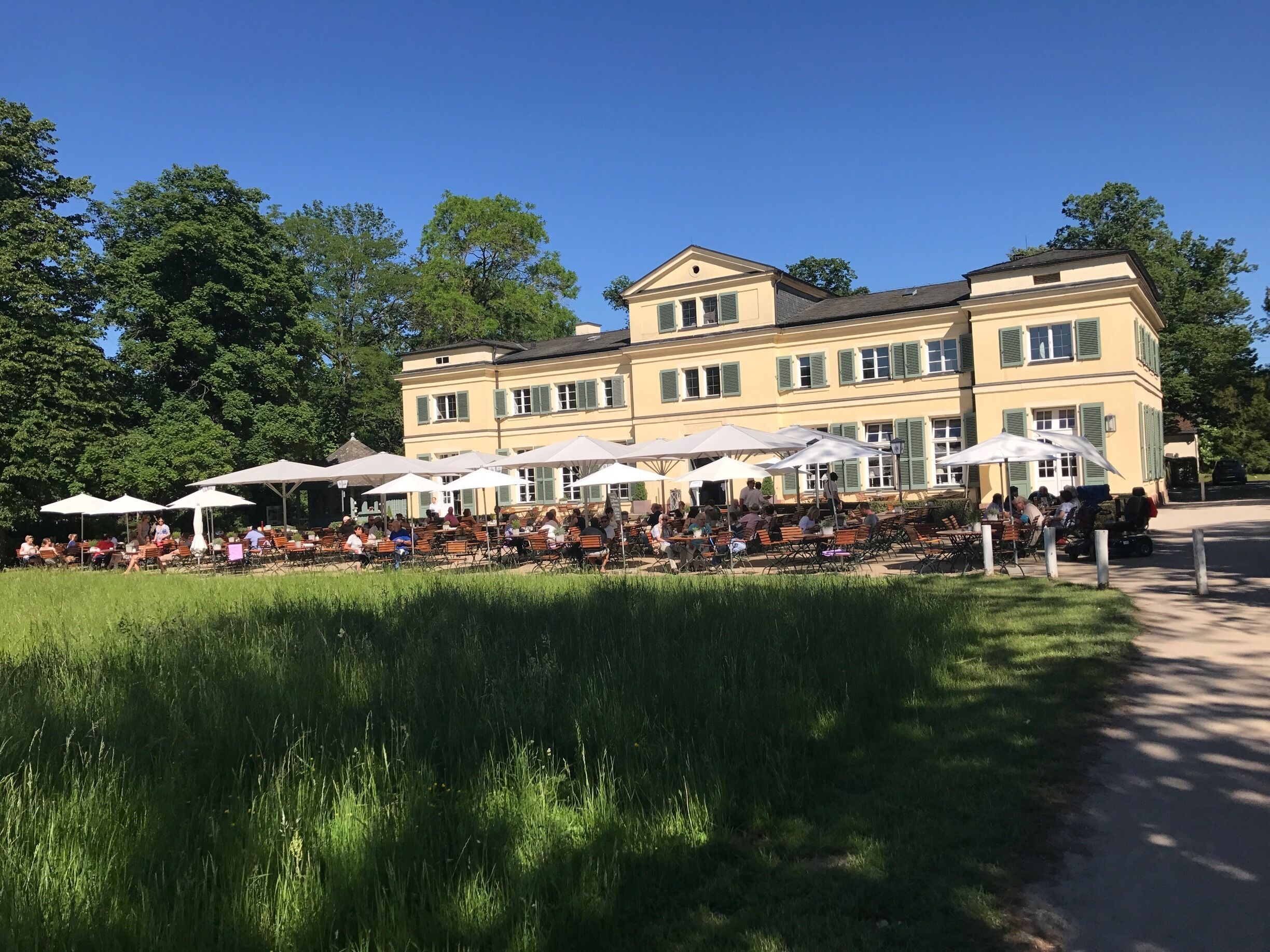A large building with outdoor seating and umbrellas, surrounded by trees and a grassy area.