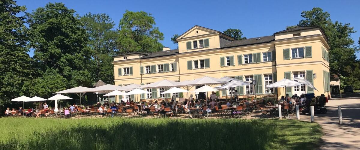 A large building with outdoor seating and umbrellas, surrounded by trees and a grassy area.