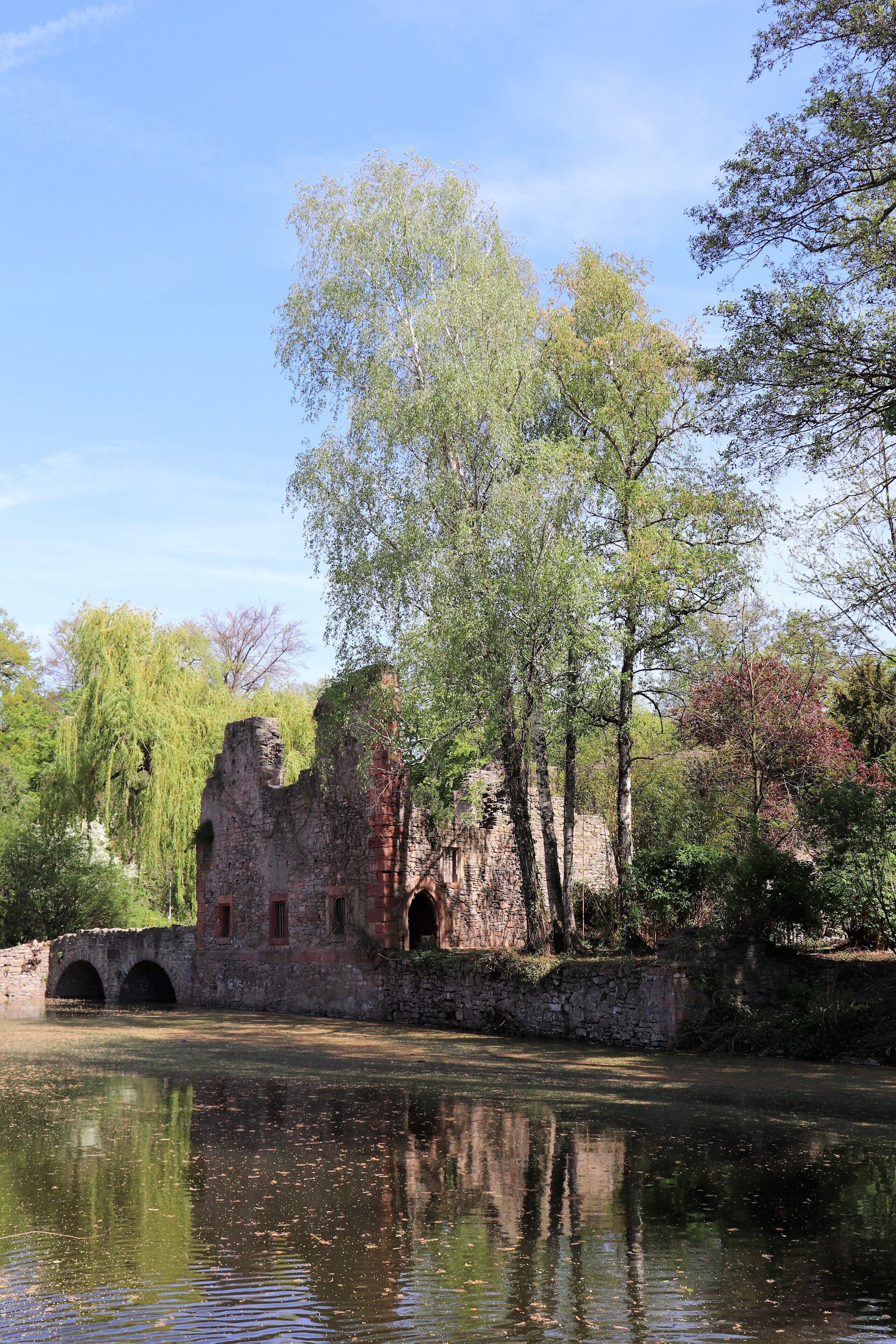 A historic stone bridge over a calm river, surrounded by lush trees and a clear sky.