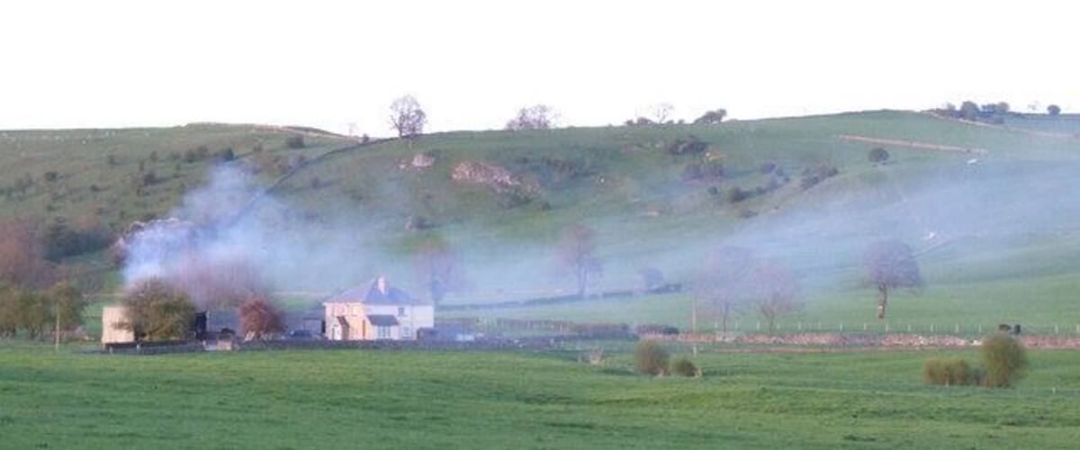 A rural landscape with a house, green fields, and smoke rising from a chimney.