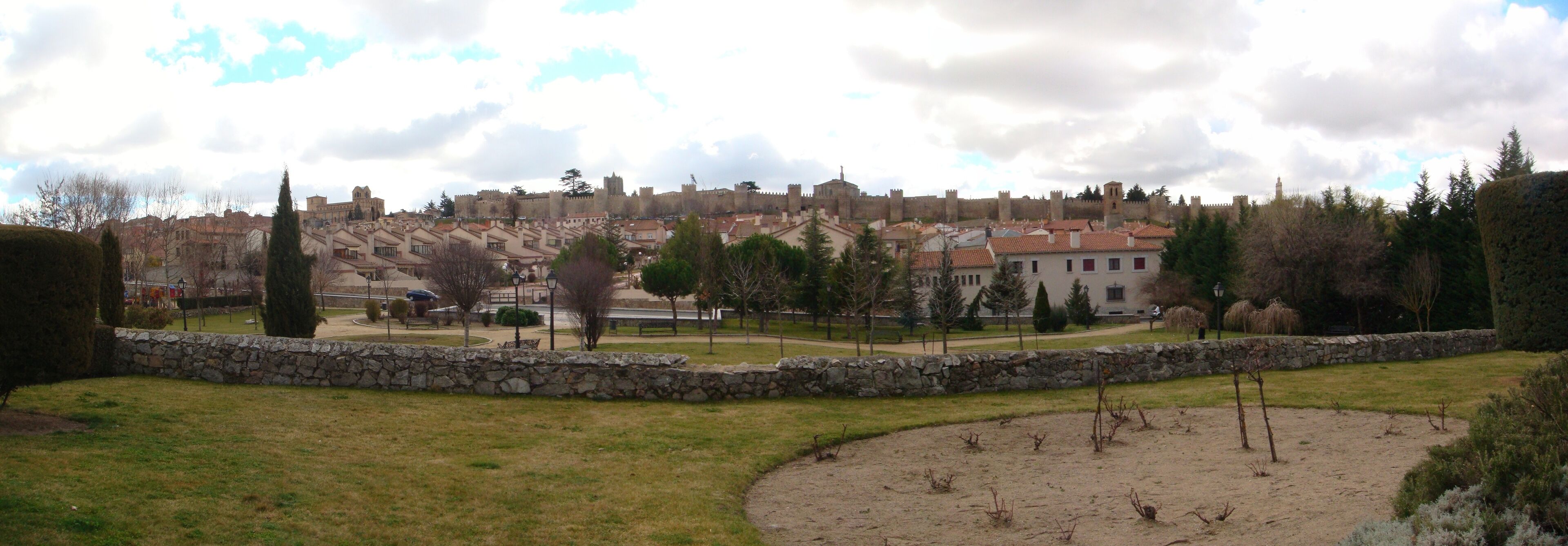A historic cityscape with medieval walls and buildings, a park with trees and a stone wall, and a clear sky.