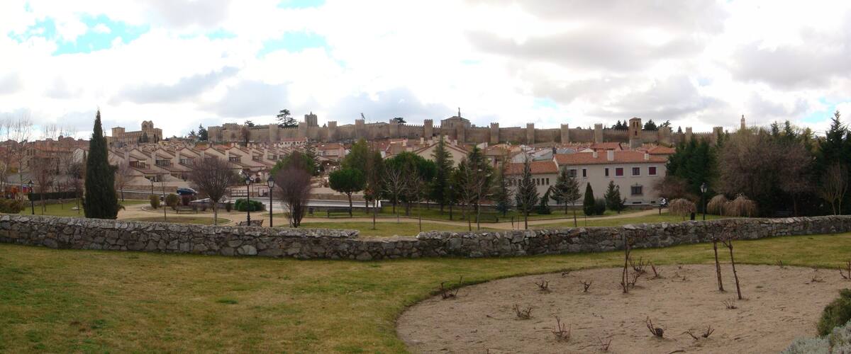 A historic cityscape with medieval walls and buildings, a park with trees and a stone wall, and a clear sky.