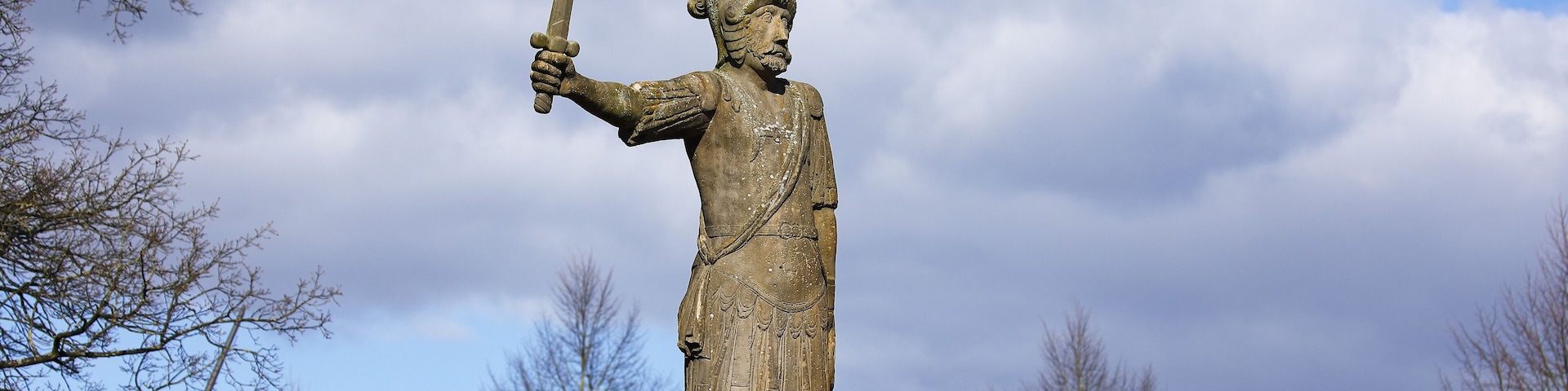 A statue of a historical figure holding a sword, set against a backdrop of red-roofed buildings and a cloudy sky.
