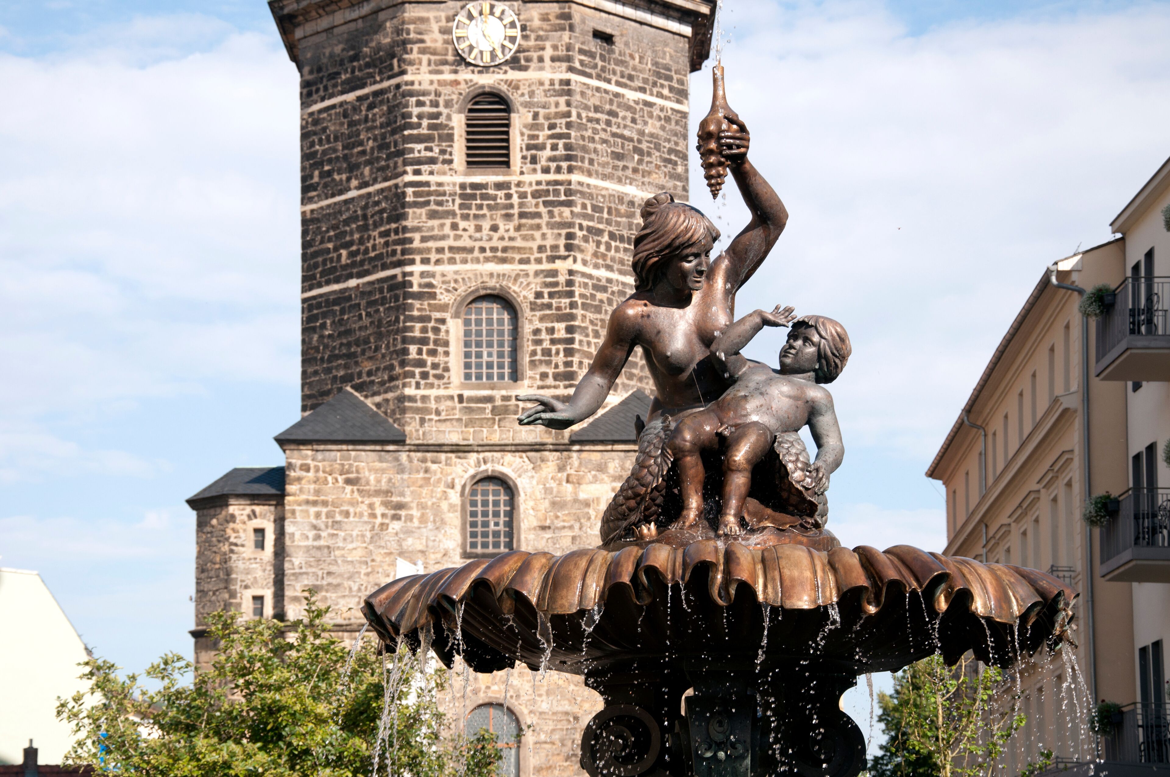 A bronze statue of a woman holding a fish, a child, and a bottle, with water flowing from the statue's base, set against a historic stone tower with a clock.