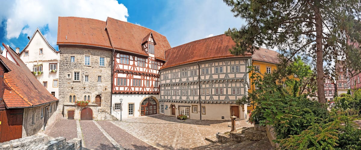 A cobblestone square with traditional half-timbered buildings, a stone building with a red roof, and a tree in the foreground.