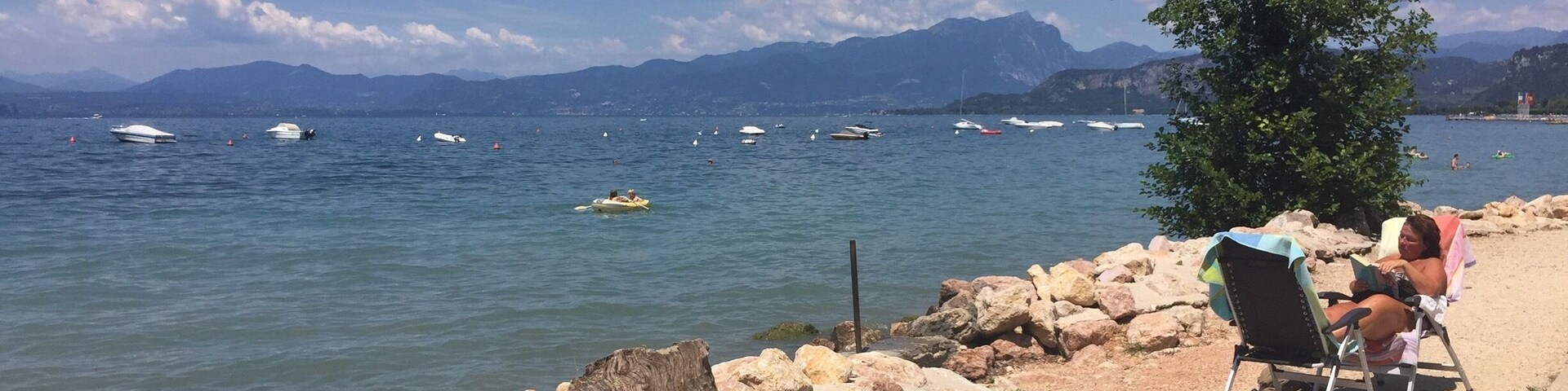 A lakeside view with boats, a rocky shoreline, and mountains in the background.