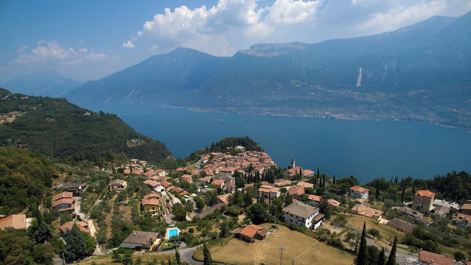 A coastal village with terraced houses, a clear blue lake, and mountains in the background.