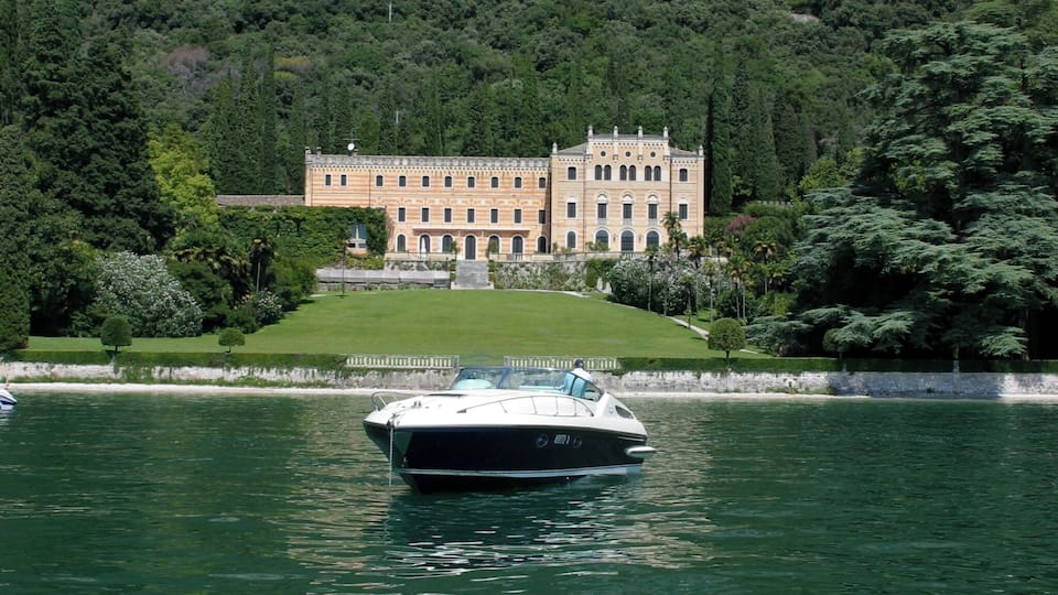 A boat on a lake with a large building surrounded by greenery and mountains in the background.