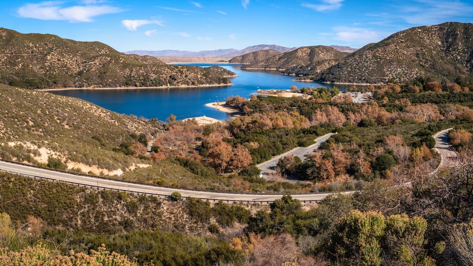 Paisaje montañoso con un camino sinuoso, un lago y árboles de colores otoñales.