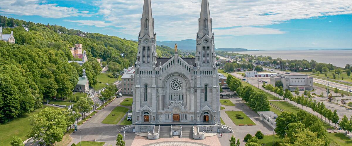 A large cathedral with two tall spires, surrounded by lush greenery and a body of water in the background.