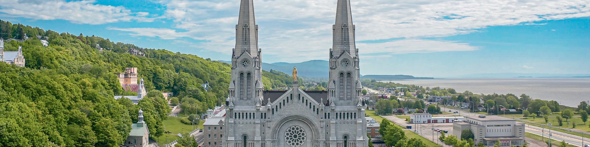 A large cathedral with two tall spires, surrounded by lush greenery and a body of water in the background.