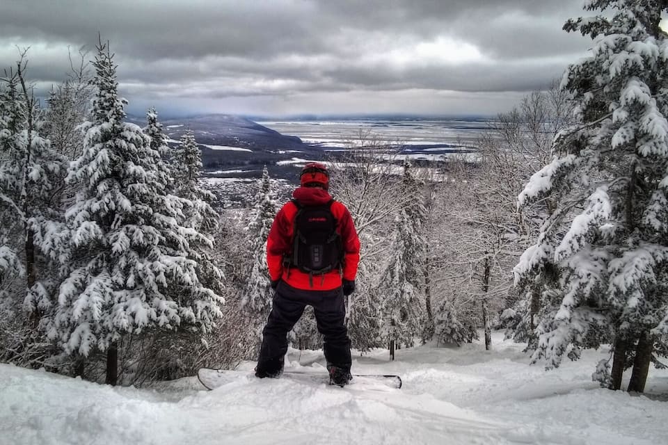 A person in a red jacket and black pants standing on a snowy slope with a backpack, overlooking a snow-covered landscape with trees and a distant view of a valley.