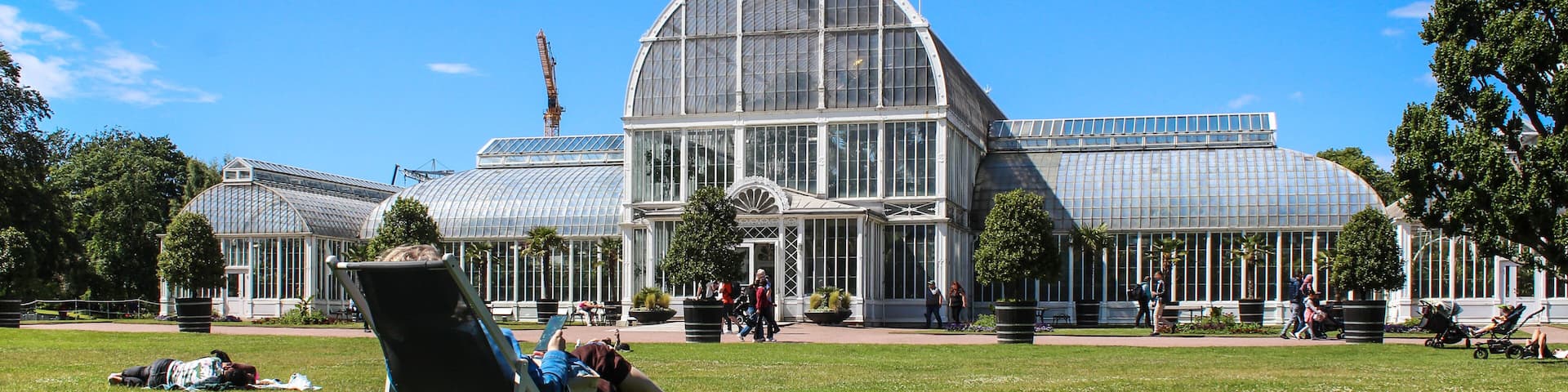 A large greenhouse with a glass dome roof, surrounded by a grassy park area with people walking and sitting on lawn chairs.