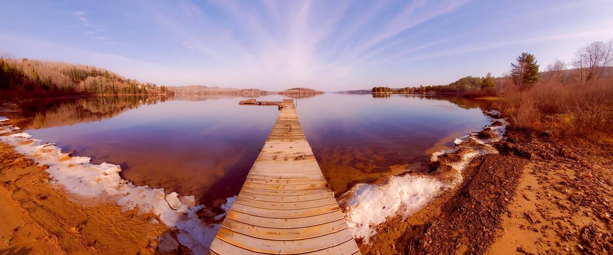 A wooden pier extending into a calm lake surrounded by trees and a clear sky.