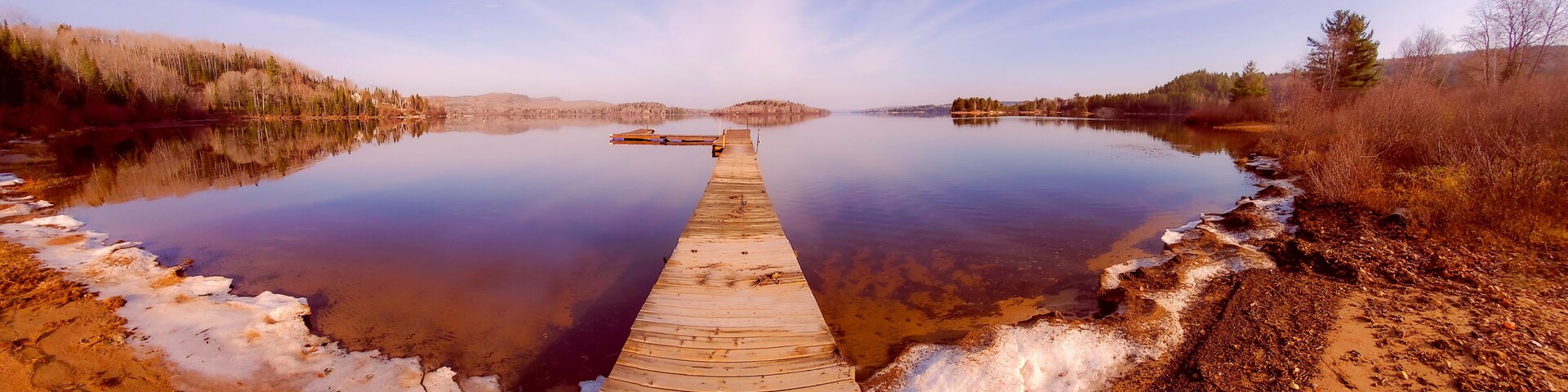 A wooden pier extending into a calm lake surrounded by trees and a clear sky.
