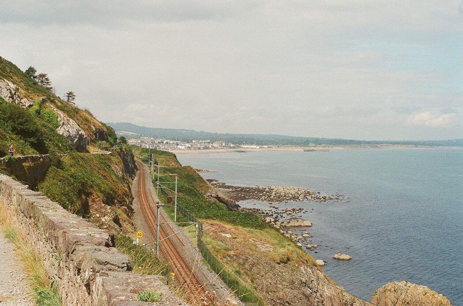 A coastal railway track with a rocky cliff, a beach, and a distant town.