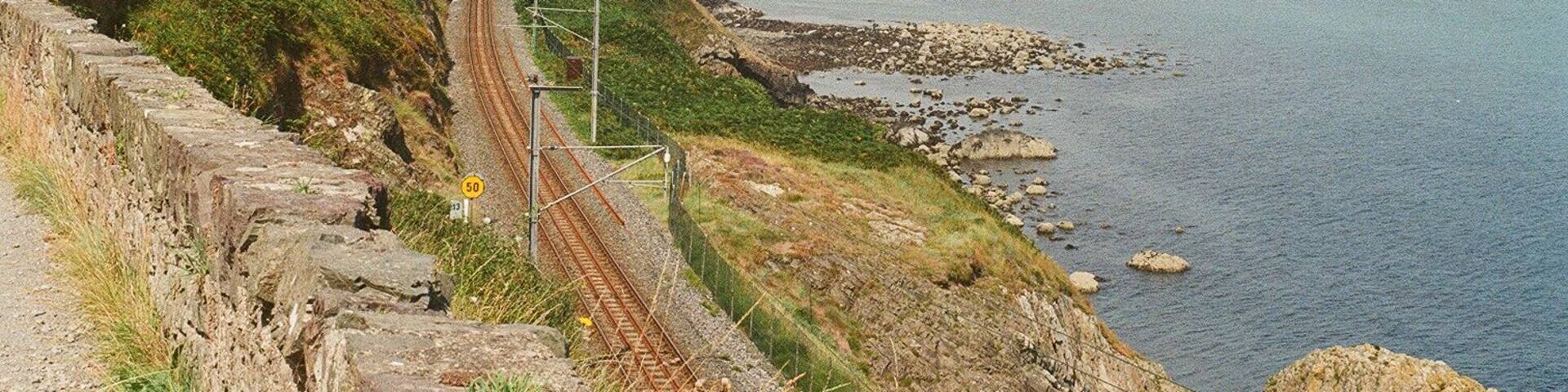 A coastal railway track with a rocky cliff, a beach, and a distant town.