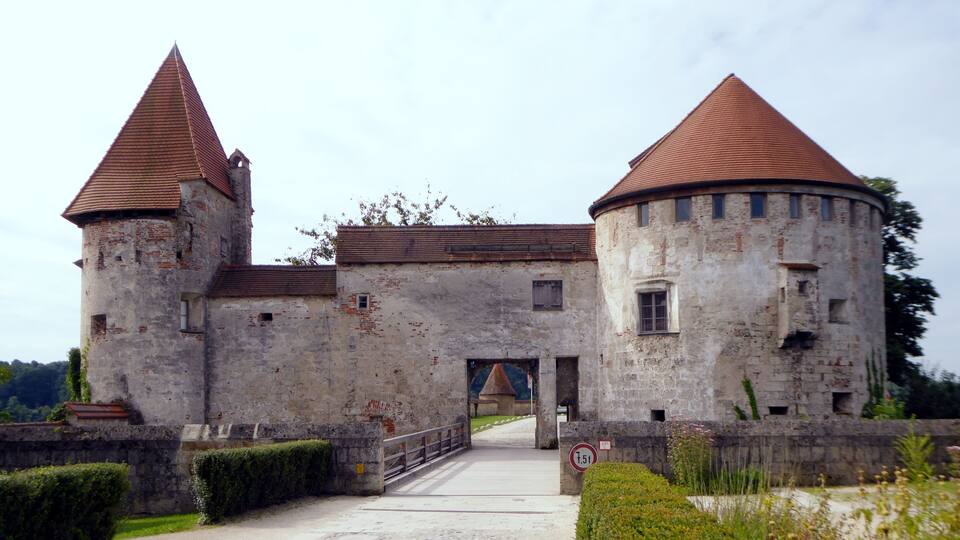 A historic castle with two towers and a round tower, surrounded by greenery.