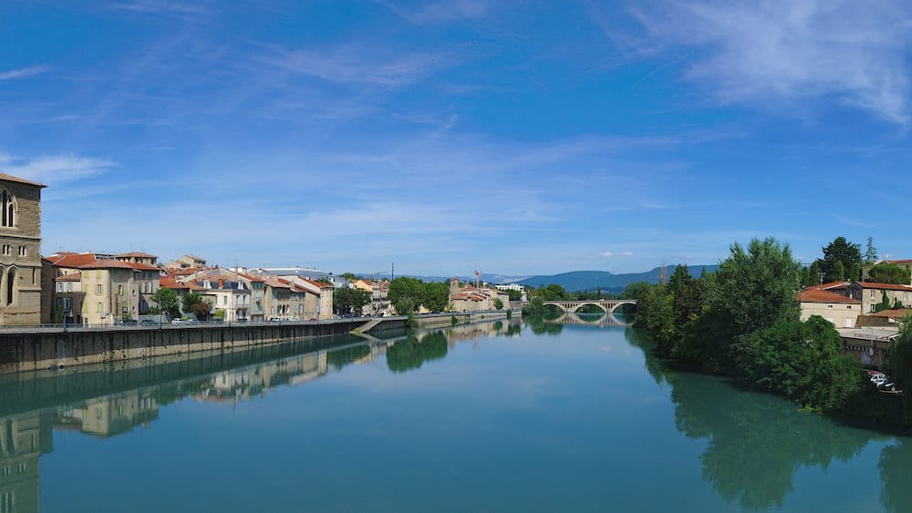 Une rivière bordée de bâtiments des deux côtés, un pont et un panneau indiquant « ROMANS » et « POURCE DE PÉAGE ».