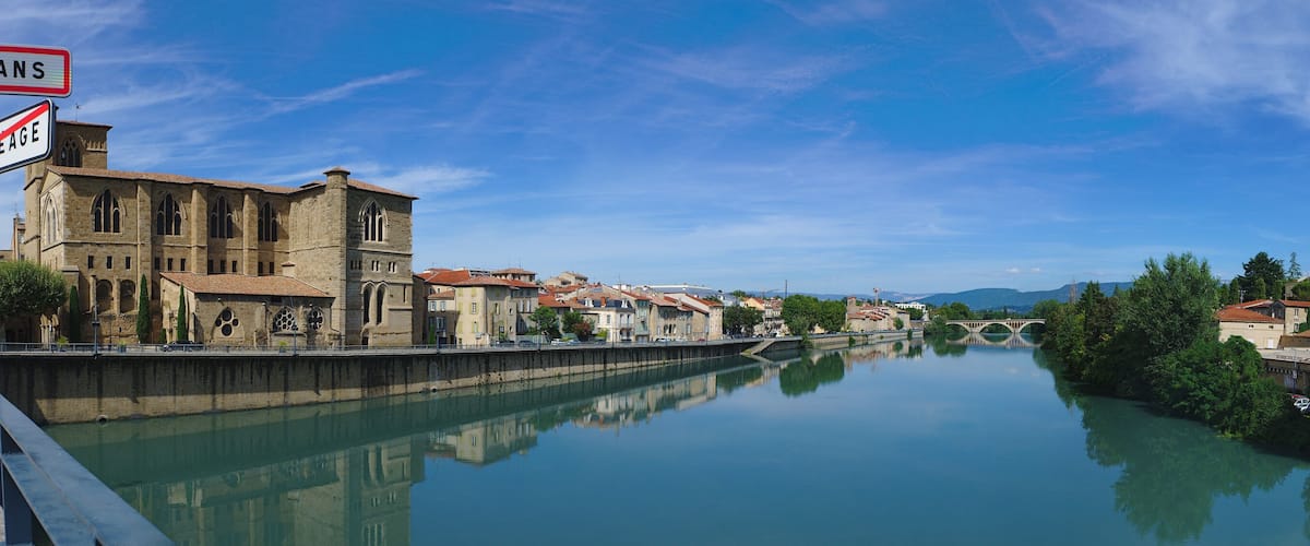 A river with buildings on both sides, a bridge, and a sign with "ROMANS" and "POURCE DE PEAGE".