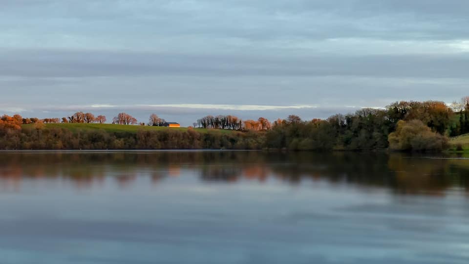 A calm lake with a forested hillside and a small building in the distance.
