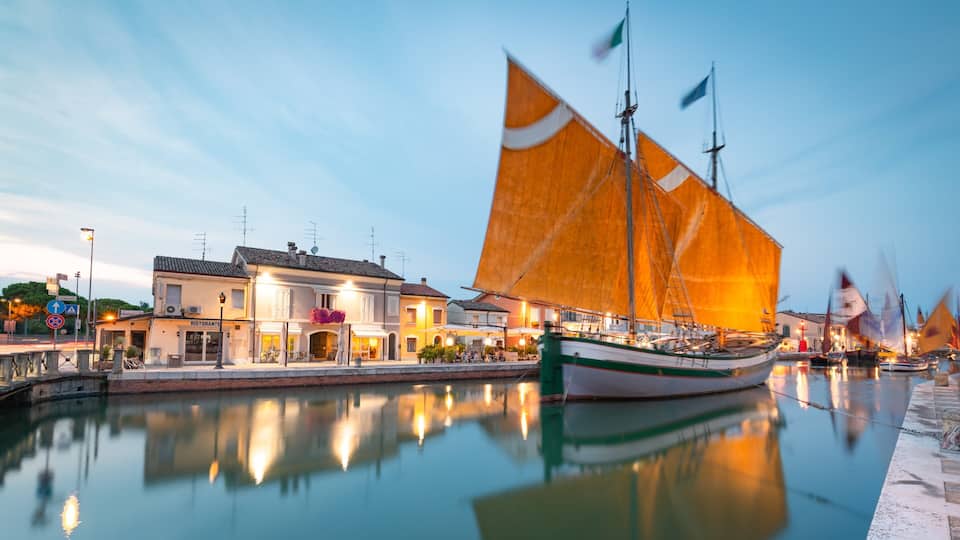A waterfront with a sailing boat, buildings, and a calm river reflecting the scene.