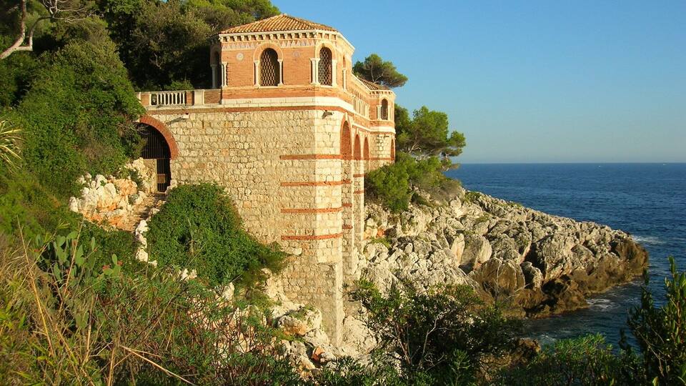 A coastal stone tower with arched windows, surrounded by rocky terrain and dense greenery.