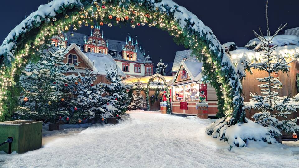 A snow-covered village with a Christmas market, decorated trees, and a traditional building with a pointed roof.