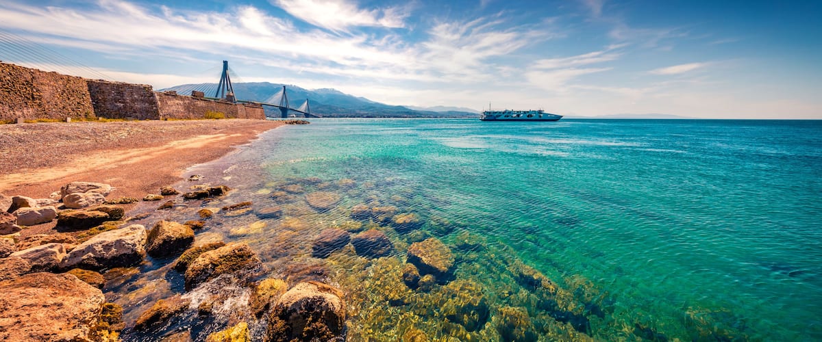 A coastal landscape with a clear blue sea, rocky shoreline, a bridge, and a boat.