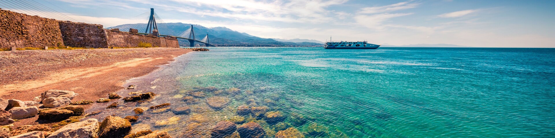 A coastal landscape with a clear blue sea, rocky shoreline, a bridge, and a boat.