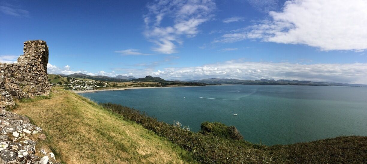 A coastal landscape with a stone ruin, grassy hills, and a calm sea under a clear sky.