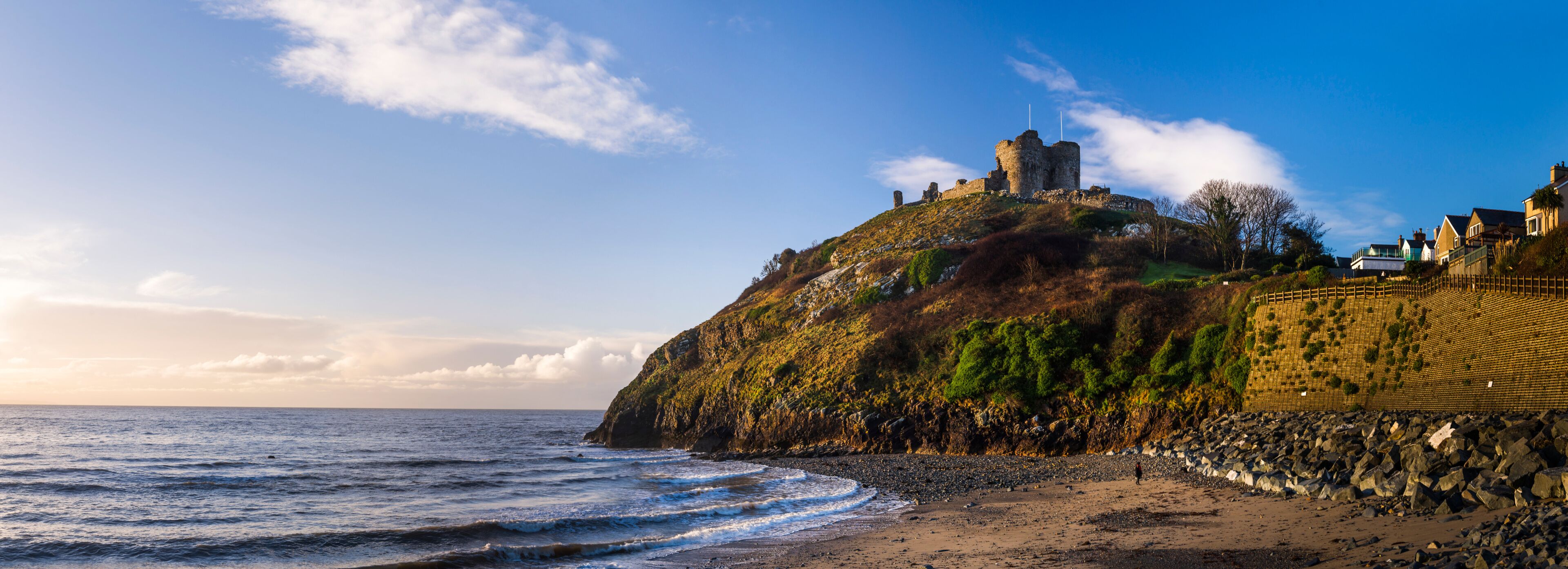 A coastal landscape with a castle on a hill, a beach, and a stone wall.