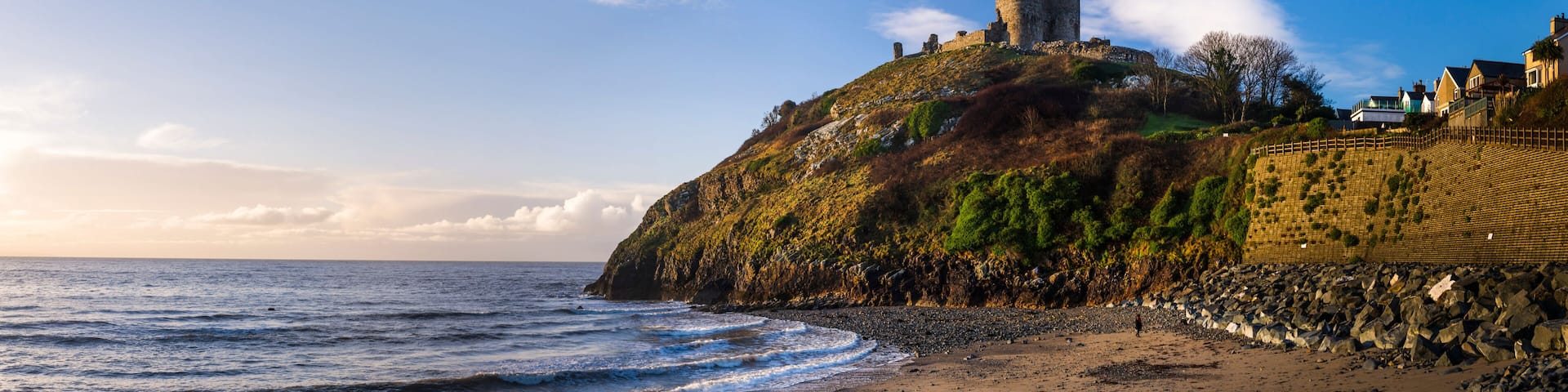 A coastal landscape with a castle on a hill, a beach, and a stone wall.