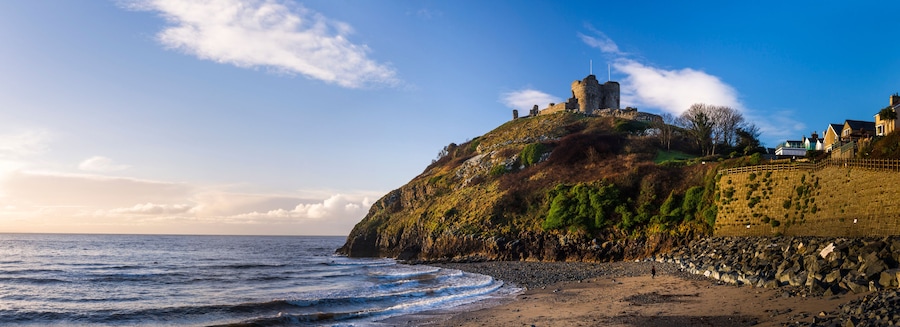 Paisagem costeira com um castelo no topo de uma colina, uma praia e um muro de pedra.