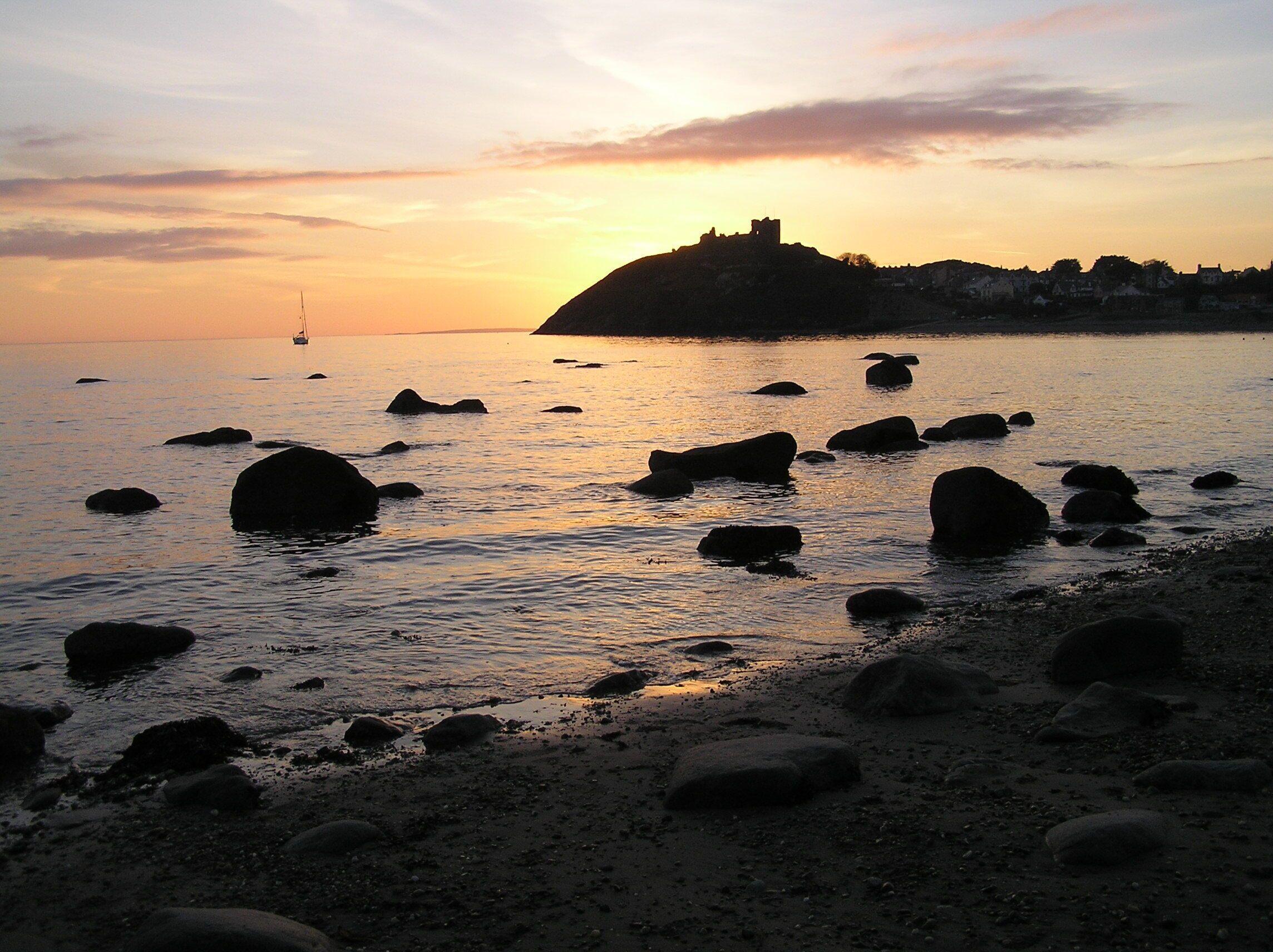 A sunset over a rocky shoreline with scattered boulders and a distant castle on a hill.