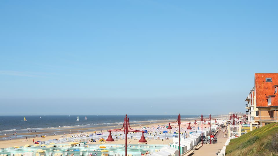 A beach with striped umbrellas and tents, a boardwalk, and a red building.
