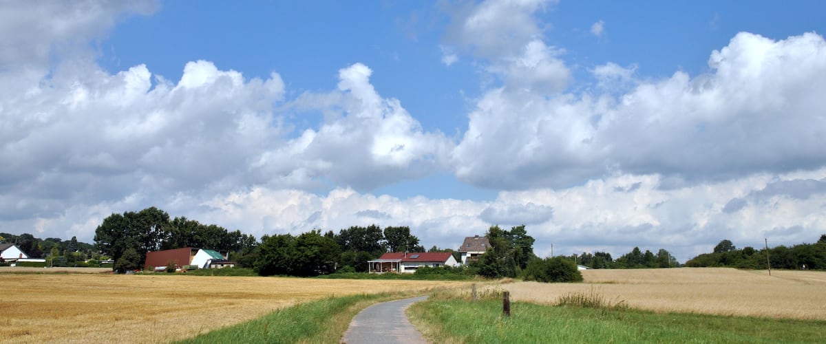 A rural landscape with a pathway leading through a field, surrounded by houses and trees under a partly cloudy sky.