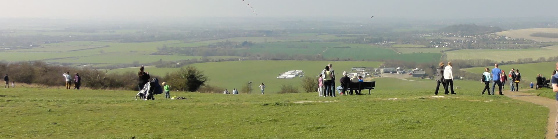 A grassy hill with people flying kites and a distant village.