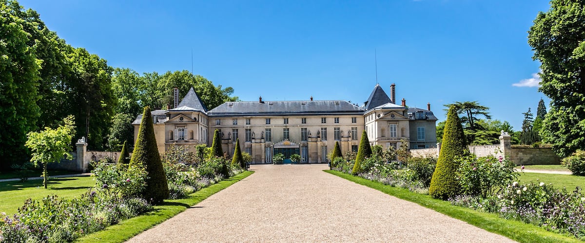 A large, historic building with a symmetrical facade, a gravel driveway, and well-manicured gardens.