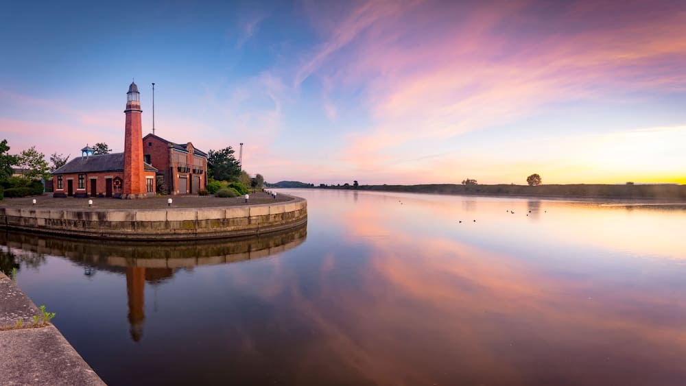 Une scène paisible au bord de la rivière, avec un bâtiment en briques doté d’une tour, entouré d’une eau calme qui reflète le ciel.