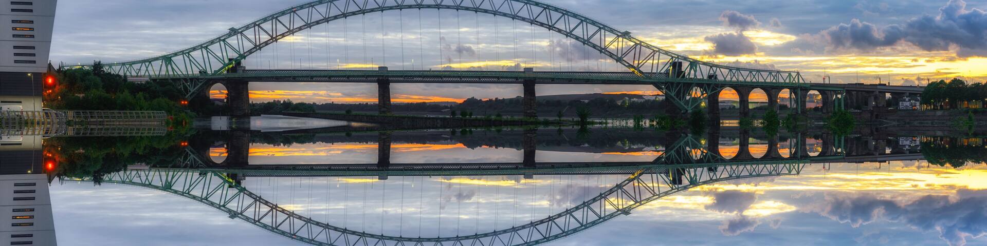 A large suspension bridge with a clear reflection in the water below, surrounded by clouds and a sunset sky.