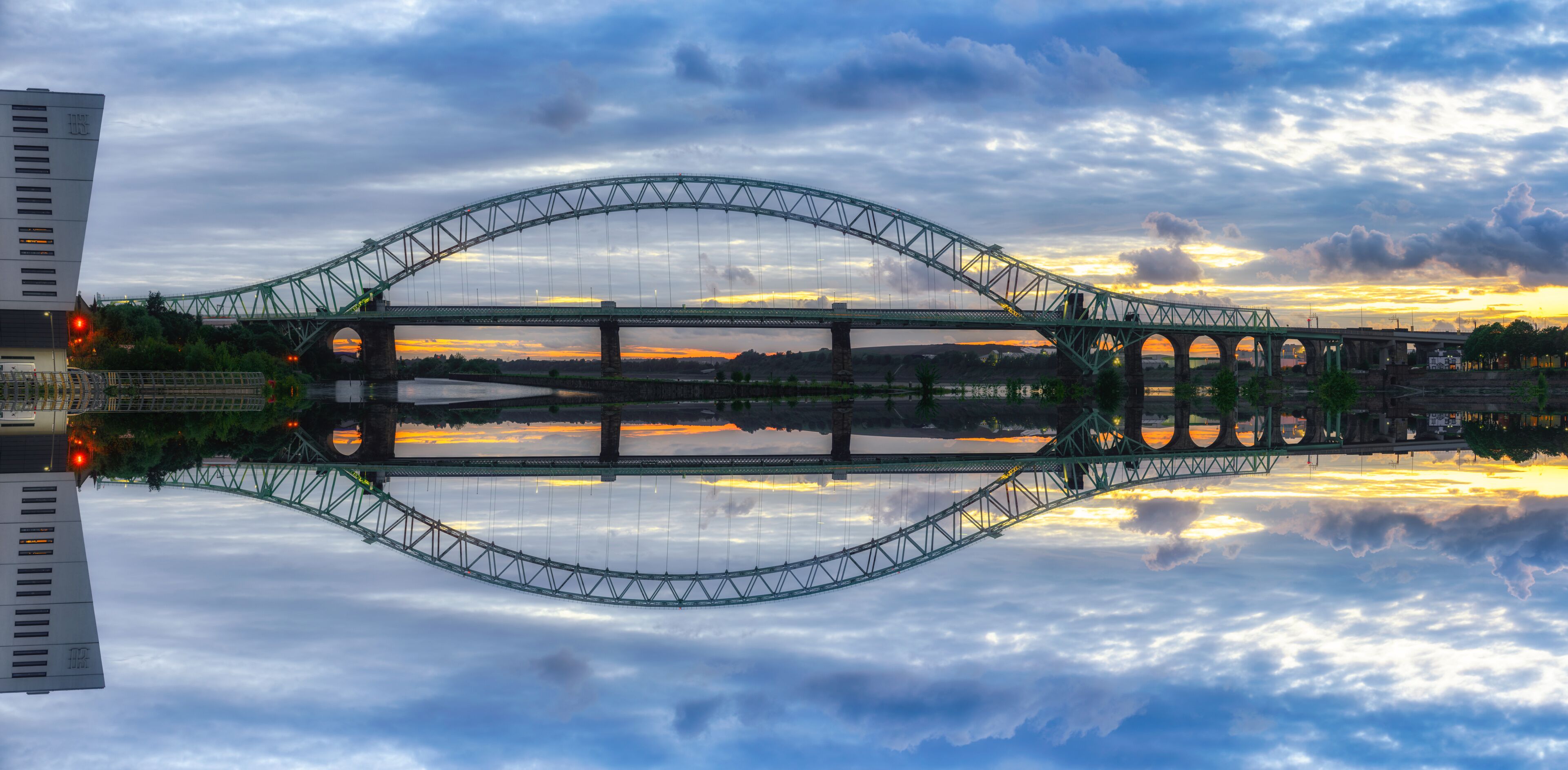 A large suspension bridge with a clear reflection in the water below, surrounded by clouds and a sunset sky.