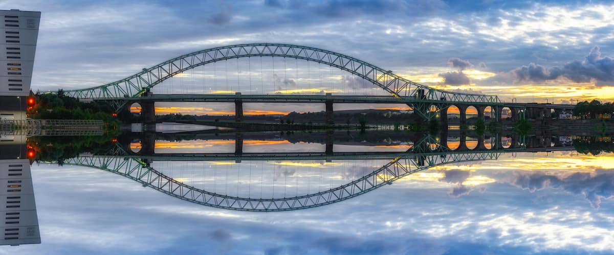 A large suspension bridge with a clear reflection in the water below, surrounded by clouds and a sunset sky.