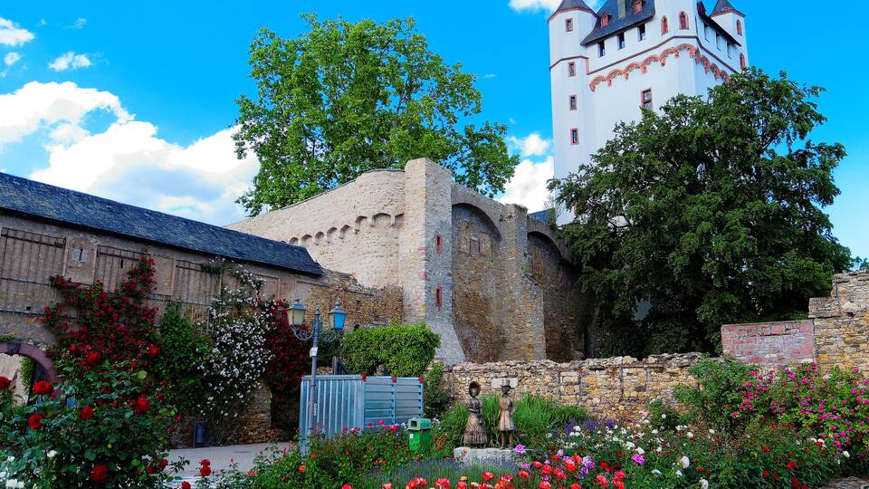 Majesteettinen, historiallinen linna, jossa on korkea torni, ja sen ympärillä on vehreitä puutarhoja ja kukkivia kasveja.