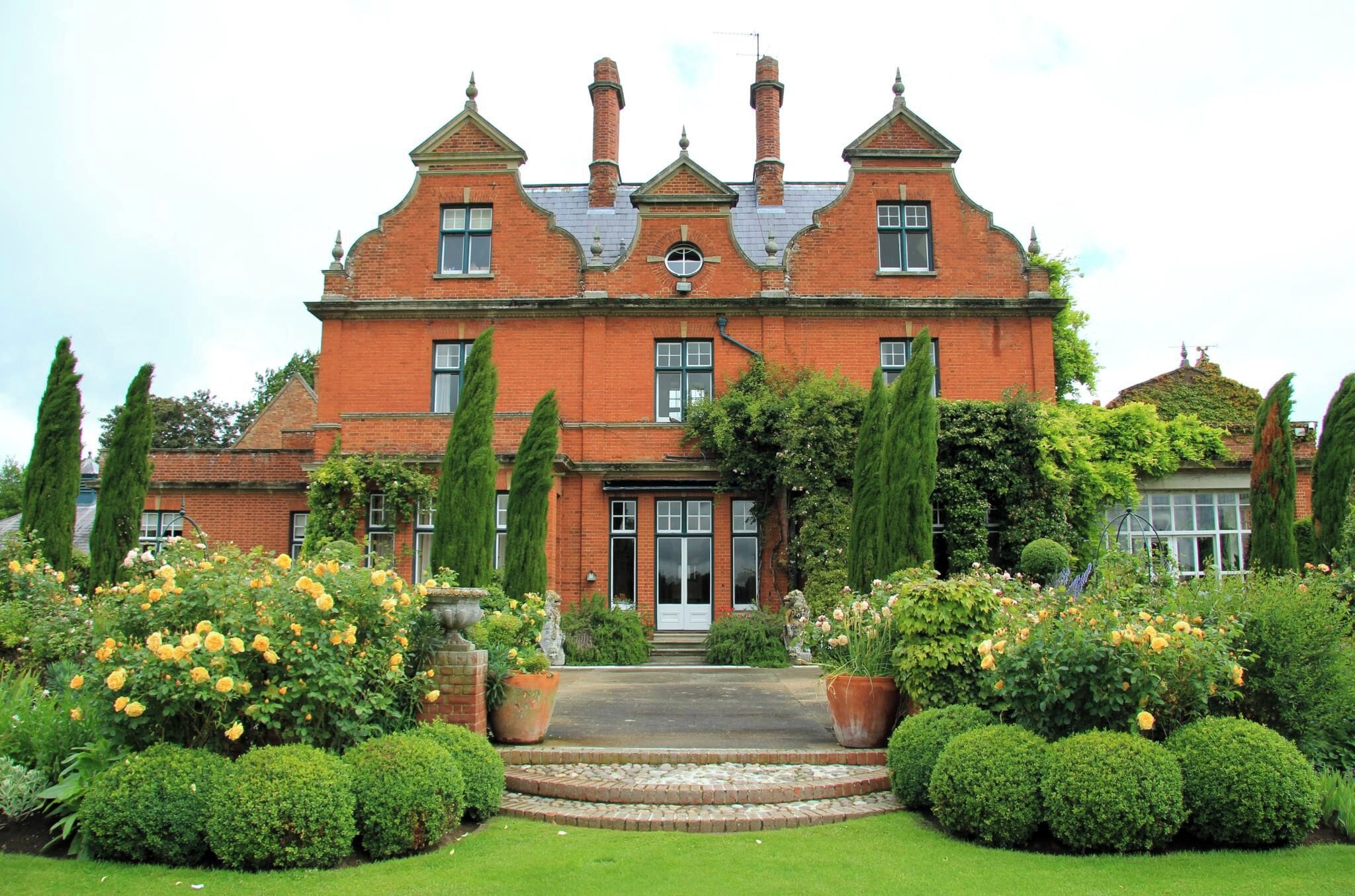A large brick house with a symmetrical facade, multiple chimneys, and a well-manicured garden with roses and trimmed hedges.